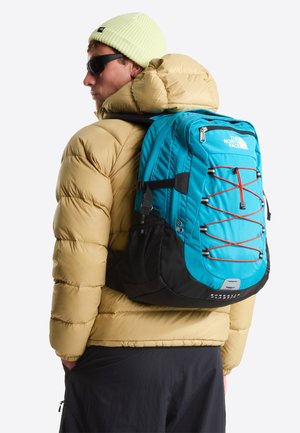 Niebiesko-czarny plecak z czerwonymi akcentami, wykonany z trwałej tkaniny. Posiada elastyczną linkę i siatkową boczną kieszeń, noszony na kurtce puchowej.