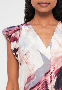 Daudzkrāsaina blūze ar V-veida izgriezumu, uzpūstām pleciem un mīkstu, viegli plūstošu faktūru. Izņēmuma ietver abstraktus rakstus sarkanu un pelēku toņu toņos.