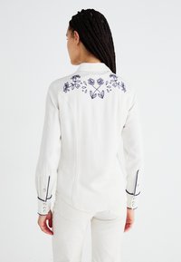 Hvit skjorte med lange ermer som har blå blomsterbroderi på ryggen, kontrastpiping langs ermene og knepping ved mansjettene.