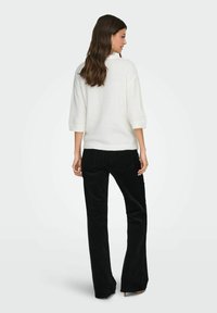 Pull en tricot crème avec manches trois-quarts, associé à un pantalon évasé en velours côtelé noir. La tenue se caractérise par un design décontracté et une texture douce.