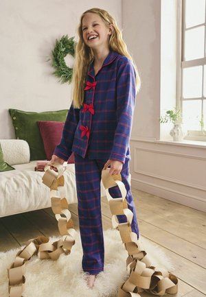 Blau karierter Pyjama-Anzug mit roten Akzenten, bestehend aus einem knöpfbaren Oberteil und passenden Hosen. Hält eine Papiergirlande. Holzboden und weicher Teppich.