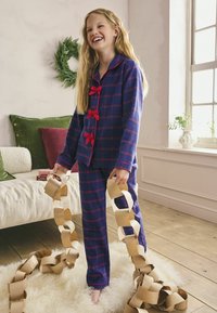 Modrý kostkovaný pyžamový set s červenými akcenty, obsahující košili na knoflíky a odpovídající kalhoty. Drží řetězovou dekoraci z papíru. Dřevěná podlaha a měkký kusový koberec.