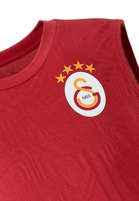 Rotes Sporttrikot mit einem strukturierten Design. Es verfügt über ein kreisförmiges Vereinslogo in Weiß, Orange und Rot, mit vier gelben Sternen darüber.