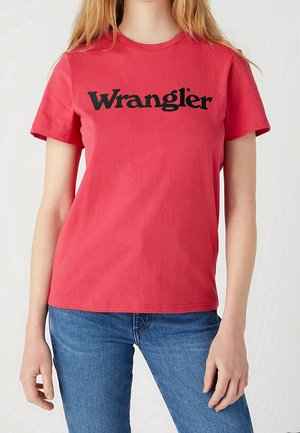 Osoba ubrana w czerwony, krótki rękaw t-shirt z napisem "Wrangler" na czarno na piersi oraz niebieskie jeansy, stojąca na jednolitym tle.