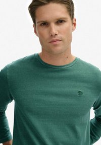 Camiseta verde de manga larga hecha de material suave, con cuello redondo y un pequeño logo bordado en el pecho. Textura suave.