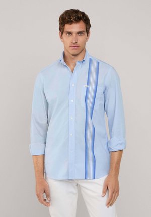 Camicia a maniche lunghe di colore azzurro chiaro con un design a righe verticali in diverse tonalità di blu, colletto con bottoni e taschino anteriore. Texture liscia.