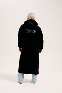Crna pufasta jakna s kapuljačom i dugom dužinom, sa srebrnim izvezenim natpisom "Juicy" na leđima, nosi se uz svijetloplave hlače.