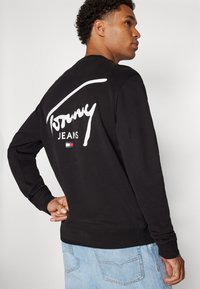 Felpa nera con logo bianco "Tommy Jeans" sulla schiena. Presenta polsini e orlo a costine. Indossata con jeans di denim azzurro chiaro.