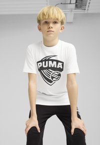 Camiseta blanca con un gran logo negro de PUMA, mangas cortas y cuello redondo, combinada con pantalones negros que presentan un detalle de logo en la pierna.