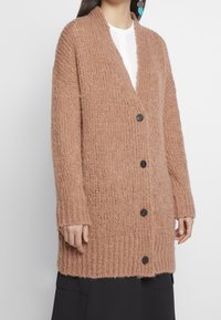 Langer, locker sitzender Cardigan aus weichem, strukturiertem beigen Strick, mit einem tiefen V-Ausschnitt, sechs schwarzen Knöpfen und gerippten Bündchen.