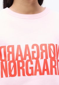 Gros plan d'une personne portant une chemise rose clair avec un grand texte rouge lisant "NØRGAARD" imprimé deux fois en style miroir.