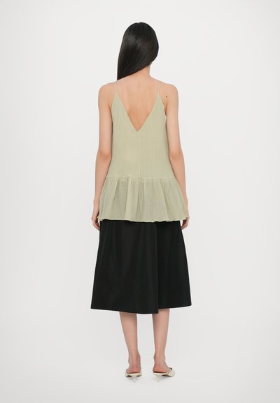 Light green, pleated tank top with a V-back design and layered hem, paired with a black, knee-length skirt and beige slide shoes.