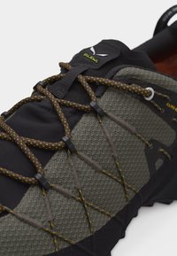 Κοντινή λήψη ενός πεζοπορικού παπουτσιού σκούρου πράσινου και μαύρου με κορδόνια κίτρινων κουκκίδων και αεριζόμενη ανώτερη επιφάνεια από πλέγμα.