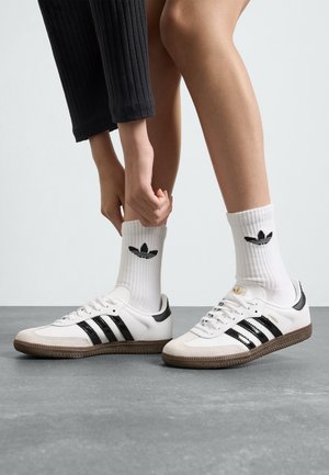 Valged Adidas Samba tossud mustade triipudega, nahast detailidega ja pruunist kummikärust. Kantud valgete soonik-sokkide kaudu, millel on must logo.