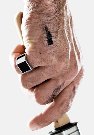 Hand som greppar ett trähandtag med en metallbas, som har en silverfärgad rektangulär ring. Handen visar svarta bläckfläckar och hårtextur.