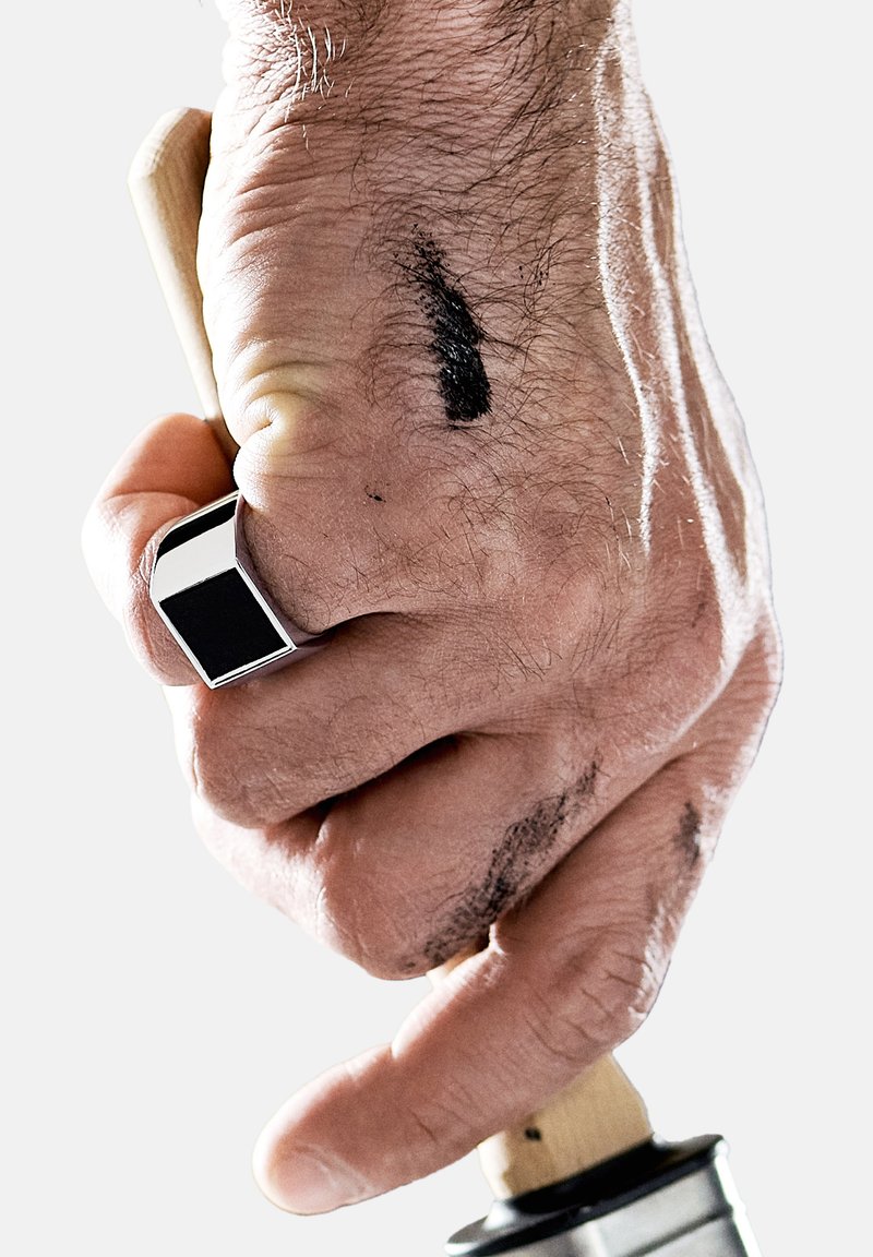 Hand som greppar ett trähandtag med en metallbas, som har en silverfärgad rektangulär ring. Handen visar svarta bläckfläckar och hårtextur.