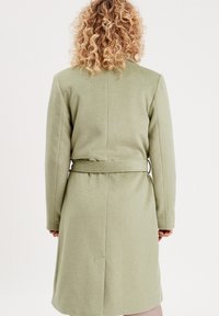 Manteau en laine vert clair, longueur genou, avec une taille ceinturée, des manches longues et une silhouette droite. Texture lisse avec un design épuré.