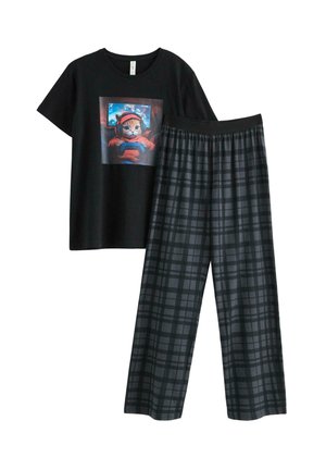 Jongen staat op blote voeten, draagt een zwart T-shirt met een kat die videospelletjes speelt en donkere geruite pyjamabroek tegen een effen achtergrond.