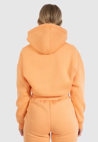 Sweat à capuche couleur pêche avec une grande capuche, épaules tombantes et poignets côtelés. Tissu doux et coupe décontractée, associé à un pantalon de survêtement assorti.