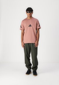 Rozā īsām piedurknēm t-krekls ar melnu Adidas logotipu un akcentiem, kombinēts ar tumši zaļām elastīgām biksēm un melniem apaviem.