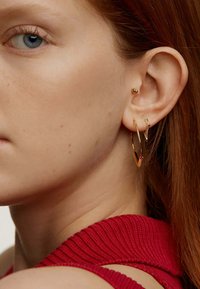 Pendientes de aro dorados y un pequeño pendiente en forma de corazón adornan la oreja, complementando un top de tela roja texturizada. La piel es clara con ligeras pecas.