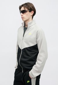 Σετ φόρμας Nike σε ανοιχτό γκρι και μαύρο με φερμουάρ μπροστά, ψηλό γιακά και λογότυπο νέον κίτρινο. Διαθέτει ελαστικά μανσέτα και ζώνη στη μέση.