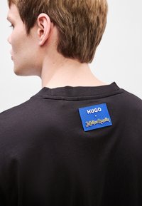 Μαύρο βαμβακερό μπλουζάκι με στρογγυλή λαιμόκοψη. Διαθέτει μπλε παραθυ ράκι με "HUGO" και "Jujutsu Kaisen" σε κίτρινο στο πίσω μέρος.