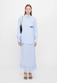 Camicia azzurra chiara con una leggera macchia viola, abbinata a una lunga gonna coordinata con un bordo in pizzo all'orlo. Accessorio borsa nera.