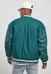 Teal bomberjack met groene leren mouwen, ribgebreide manchetten en tailleband, en witte gestreepte accenten. Glad textuur en een relaxed fit.