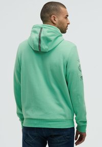 Sudadera verde menta hecha de tela suave, con un detalle de capucha a rayas negras, puños acanalados y estampados en la manga izquierda.