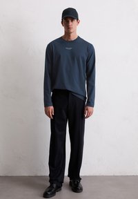 Μακρυμάνικο σκούρο μπλε πουκάμισο με στρογγυλή λαιμόκοψη και λογότυπο στο στήθος, συνδυασμένο με μαύρο παντελόνι και μαύρες παπούτσια, ολοκληρωμένο με ένα ταιριαστό καπέλο.