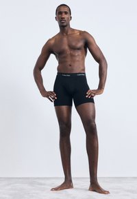 Fekete pamut boxer alsónadrág, amely illeszkedő szabású és rugalmas derékpánttal rendelkezik, finom fehér logóval. A modell mezítláb áll egy szürke felületen.