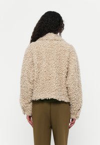 Beige fluffig jacka med en texturerad, lockig yta. Har en krage, kort design och långa ärmar. Bärs över olivgröna byxor.