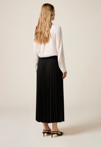 Pitkä musta laskostettu hame yhdistettynä valkoiseen puseroon, jossa on pitkät hihat ja nappiyksityiskohdat, täydentäen kokonaisuuden mustilla korkokengillä.