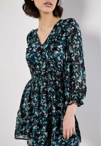 Robe florale noire avec des fleurs turquoise et blanches. Décolleté en V et taille smockée avec des manches bouffantes. Tissu léger et semi-transparent.