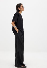 Črna majica s sproščenim krojem, v kombinaciji s širokimi hlačami z črnimi črtami. Dodatki vključujejo elegantne sončne očala in teksturiran srebrn zapestnico.