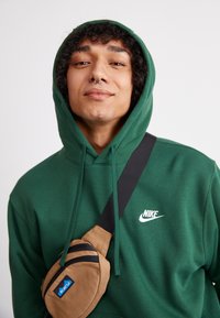 Zaļš kapuce ar baltu Nike logo, saskaņots ar brūnu Kavu krustsoma, kurai ir melni rāvējslēdzēji un siksna. Gluda auduma tekstūra.