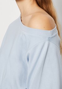 Sudadera azul claro de hombros al aire con cuello acanalado, textura suave y hombros caídos, que muestra un ajuste relajado y un diseño casual.