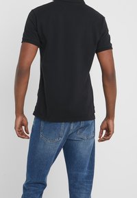 Man i kortärmad svart pikétröja och blå jeans som ses bakifrån mot en enfärgad ljus bakgrund.