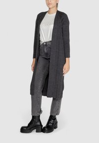 Lungo cardigan grigio aperto davanti, realizzato in materiale morbido, abbinato a una t-shirt grigio chiaro con lettering decorativo e jeans grigi a vita alta.