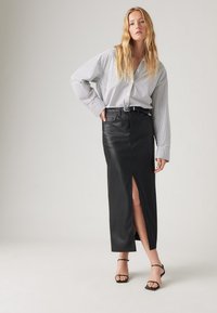 Jupe en cuir noir avec une fente frontale, assortie à une ceinture noire. Chemise à rayures avec boutons et manches larges. Modèle portant des sandales noires à lanières.