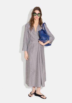 Kobieta w długiej szarej sukience w paski, duże okrągłe okulary przeciwsłoneczne, czarne sandały, trzyma dużą niebieską torebkę na lewym ramieniu.