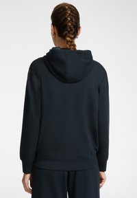 Donna con capelli intrecciati che indossa una felpa nera con cappuccio e pantaloni neri, mostrata di spalle su uno sfondo bianco uniforme.