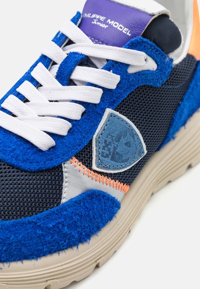 Πολύχρωπα αθλητικά παπούτσια με μπλε σουέτ επάνω μέρος, διχτυωτό ύφασμα, λευκές κορδόνες, μωβ και πορτοκαλί λεπτομέρειες, και υφασμένο μπεζ πάτο.