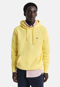 Geel hoodie met een trekkoordcapuchon, buidelzak en subtiele logo. Draag het over een roze gestreept shirt, met daaronder een marineblauwe broek zichtbaar.