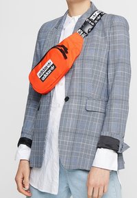 Orange Adidas crossbody bag with black logo, rectangular shape, adjustable strap featuring a patterned design, worn over a plaid blazer.