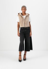 Donna con capelli biondi corti che indossa un gilet di pelliccia beige, una maglietta bianca, jeans neri a gamba larga e scarpe con cinturino alla caviglia nere, in piedi contro un muro bianco.