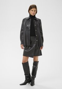 Chaqueta de camisa de cuero negro con botones a presión, combinada con una falda de cuero a juego. Botas de cuero negro hasta la rodilla completan el conjunto.