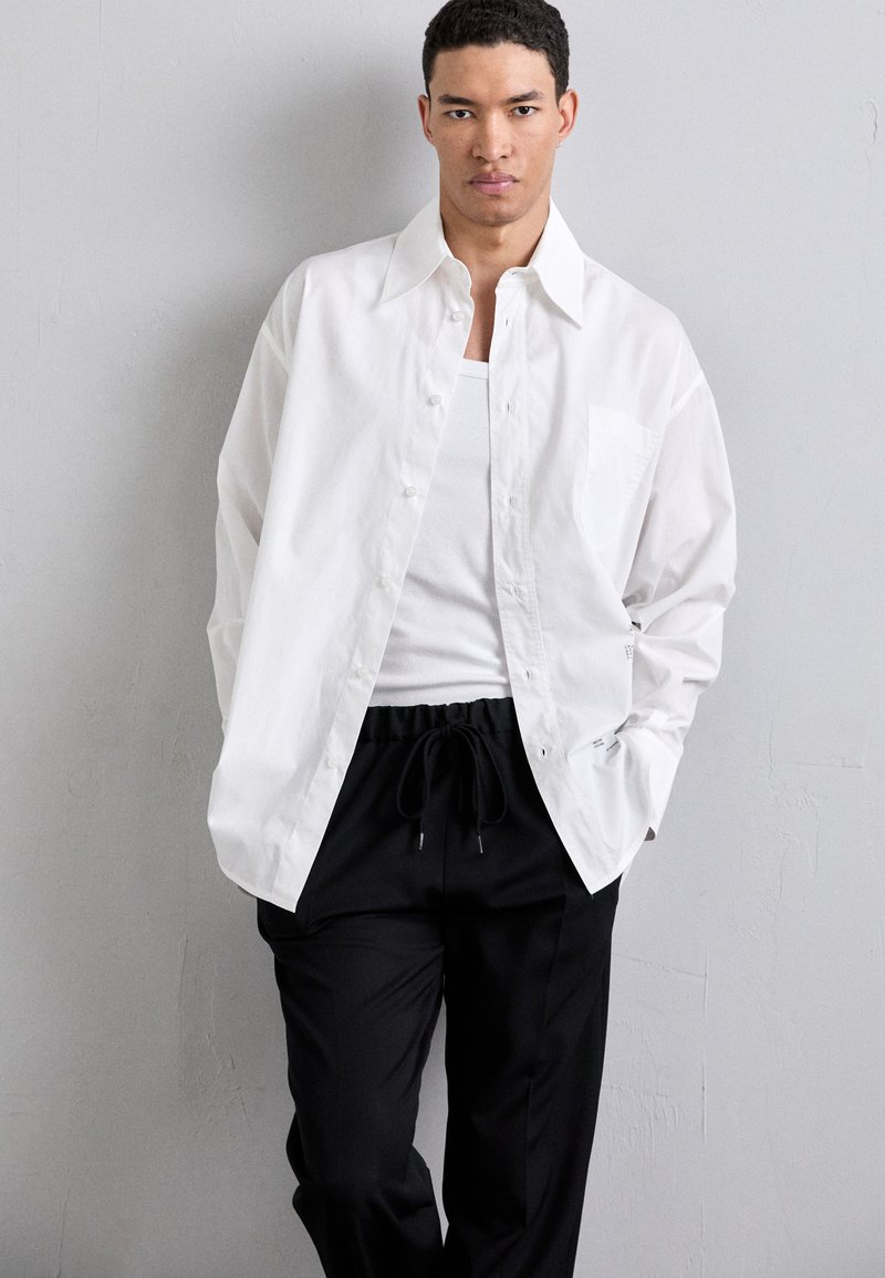 Hvid skjorte med knaplukning og krave, frontlomme og afslappet pasform, parret med sorte bukser med snøre, sat imod en neutral baggrund.