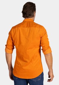 Langemouwige oranje shirt, van katoen, met een knoopsluitende kraag, opgerolde mouwen en details op de rug. Draagt met donkerblauwe jeans.